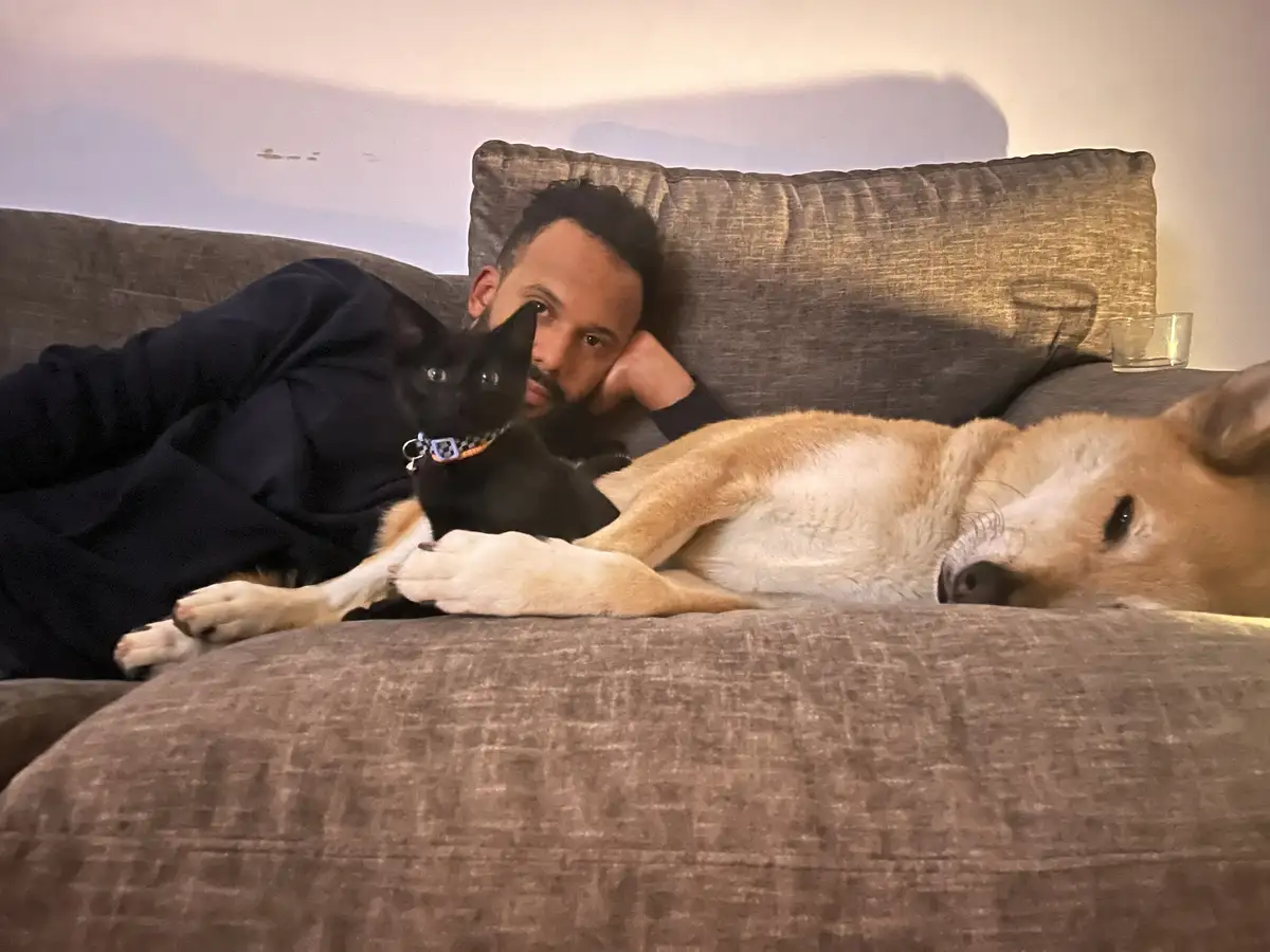 A man in a dark hoodie lies on a gray couch, propped up on one elbow, looking directly at the camera with a tired…