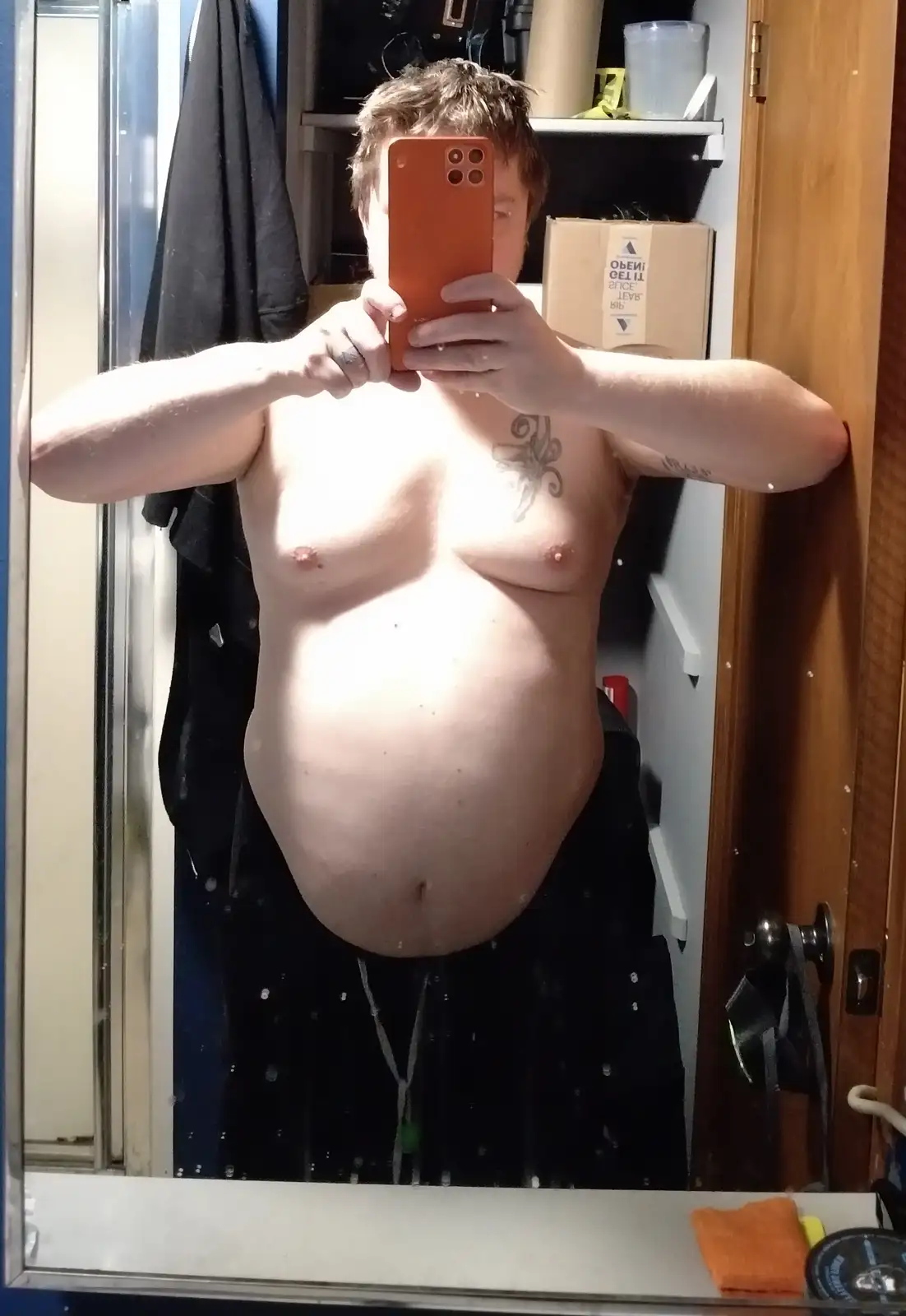 A shirtless person with a large belly takes a mirror selfie in a bathroom, holding an orange phone. The image shows tattoos…