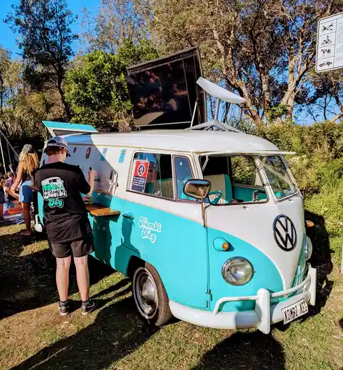 A vintage turquoise and white Volkswagen Kombi van is parked on grass with its side door open, serving drinks from a…