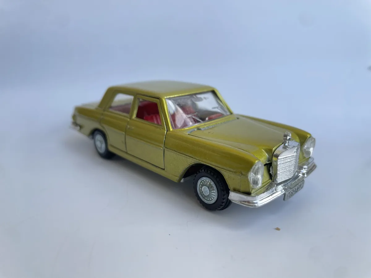 A gold-colored toy car, resembling a classic Mercedes-Benz, is photographed on a plain white surface. The car has red…