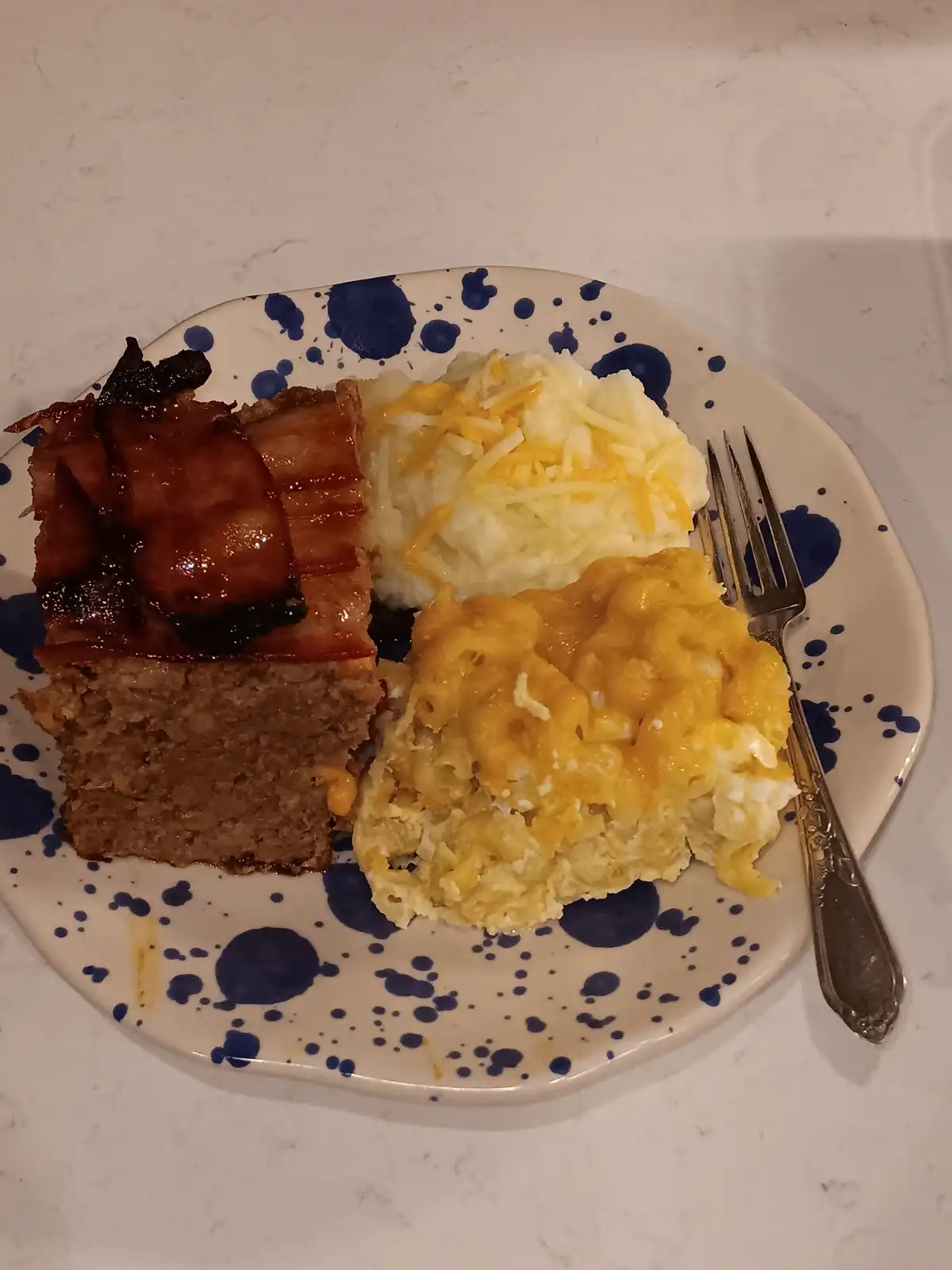 A plate of food featuring a slice of meatloaf topped with bacon, mashed potatoes with shredded cheese, and scrambled eggs…