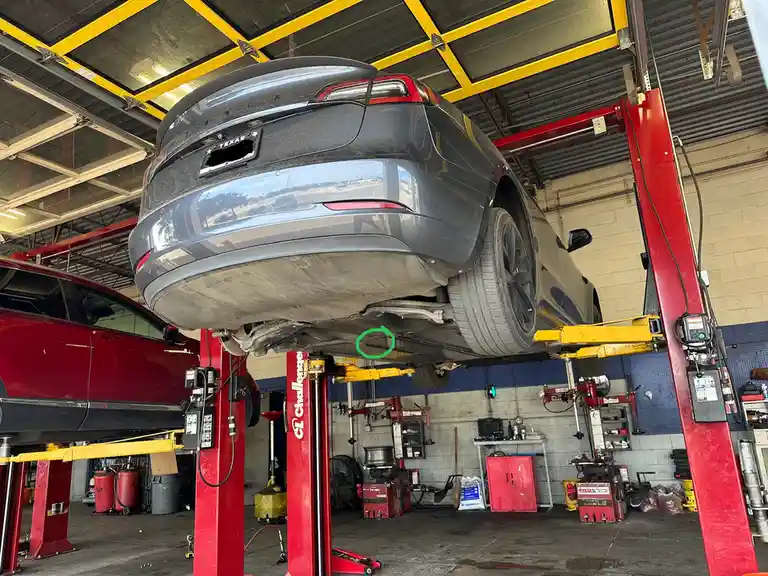 A gray Tesla Model X is suspended upside down on a red and yellow car lift in an auto repair shop, with a green circle…