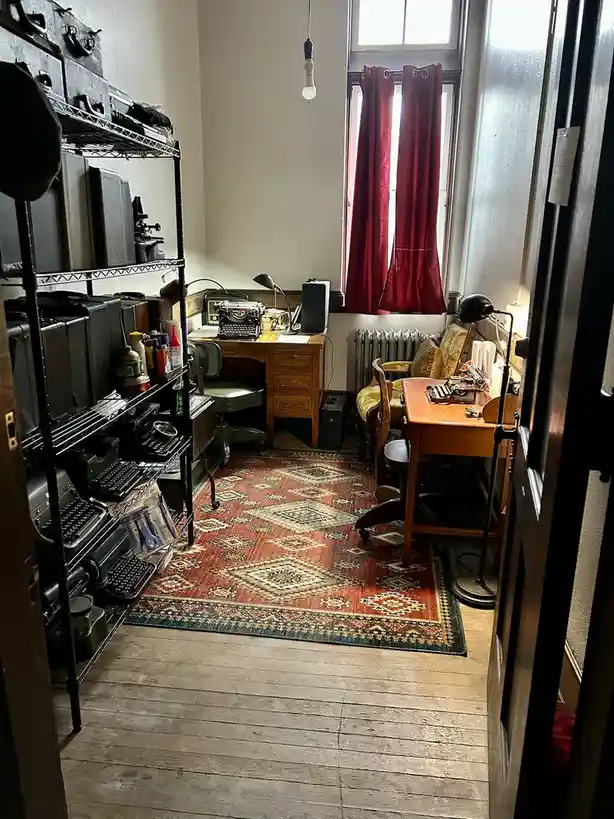 A cozy, cluttered room viewed from a doorway, featuring two wooden desks with vintage typewriters, a metal shelving unit…