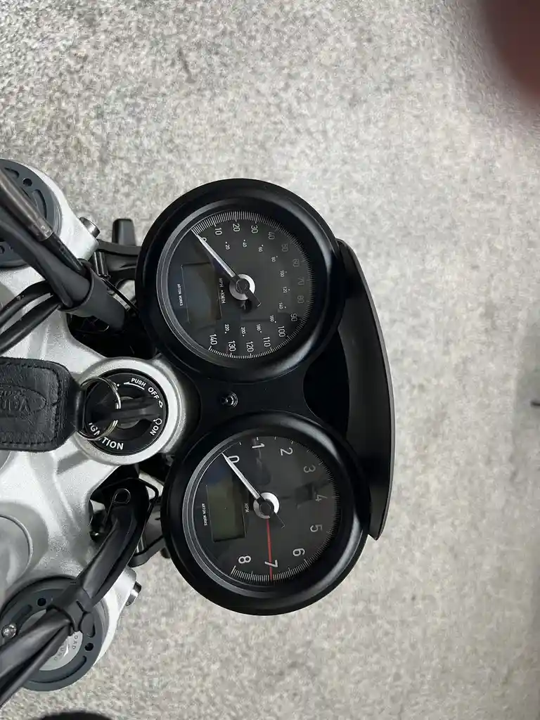 A close-up photo of a motorcycle's instrument cluster shows a speedometer and tachometer, both needles pointing to zero…
