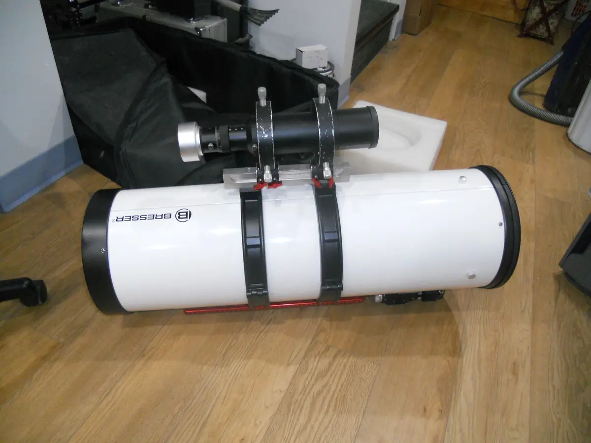A white Bresser brand telescope lies horizontally on a wooden floor, secured with black straps and mounted with a black…