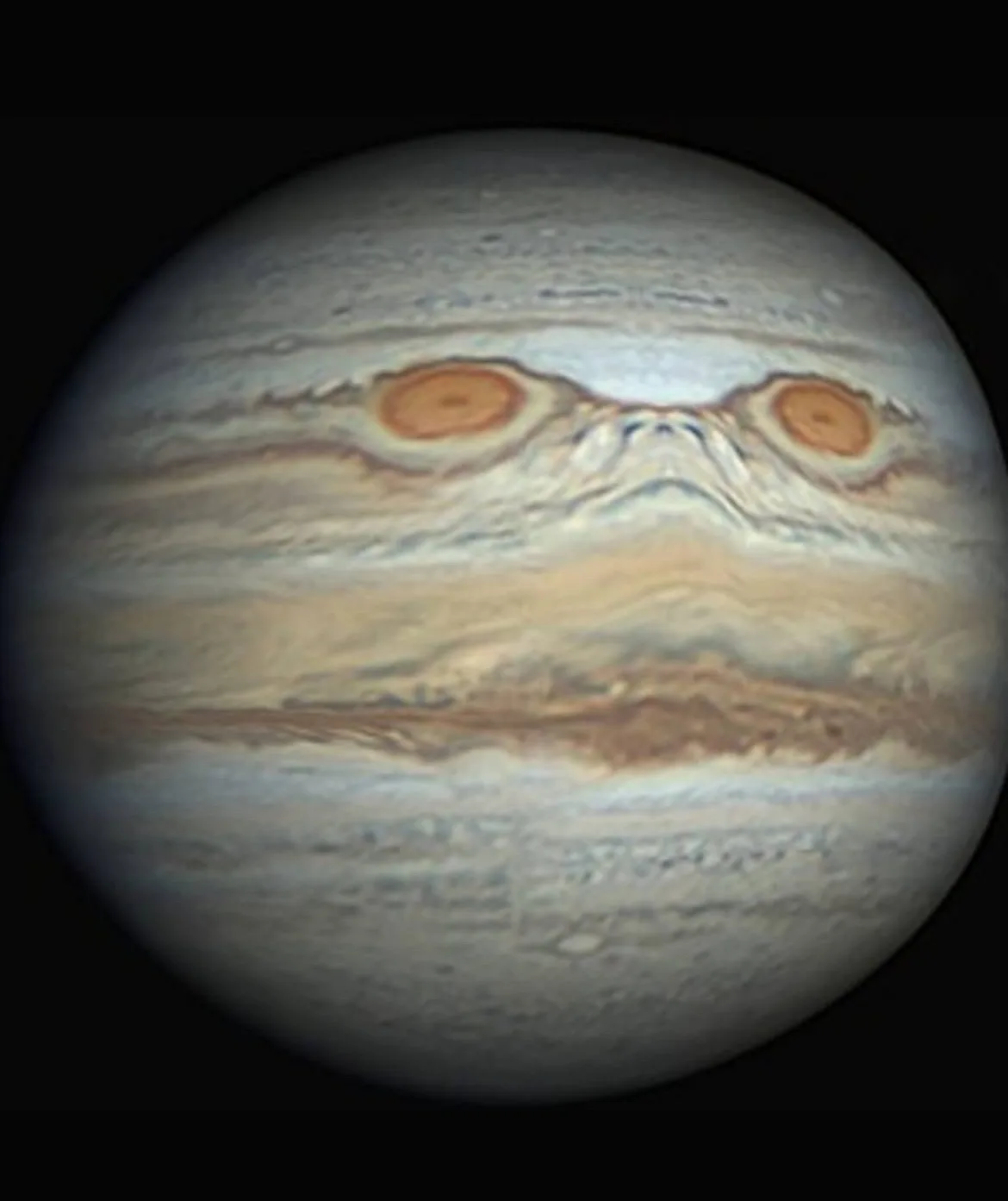 The image shows the planet Jupiter with its swirling cloud bands edited to resemble a face, featuring two large orange storm…