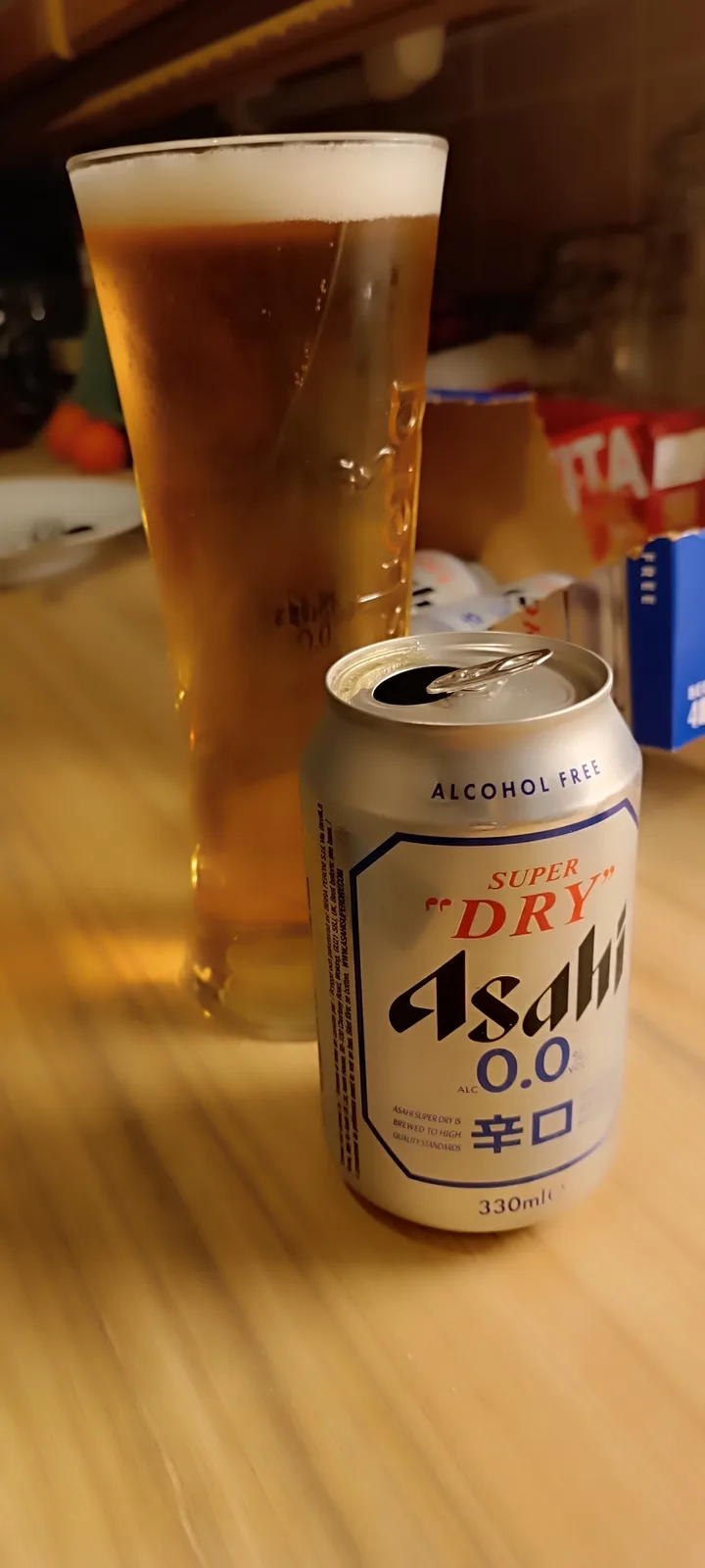 A can of Asahi Super Dry 0.0% alcohol-free beer sits open on a wooden table next to a tall glass filled with amber-colored…