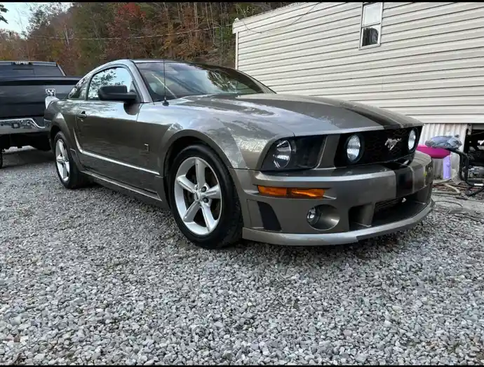 A gray Ford Mustang with black racing stripes is parked on gravel in front of a beige house, with a black pickup truck…