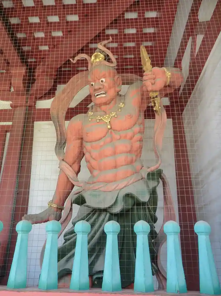 A large, muscular, red-skinned statue of a wrathful deity with a fierce expression, holding a sword, sits behind a wire mesh…