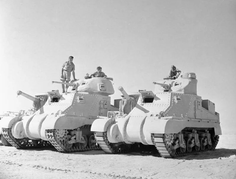 A black-and-white historical photograph shows three military tanks parked in a desert, with soldiers standing on top of…
