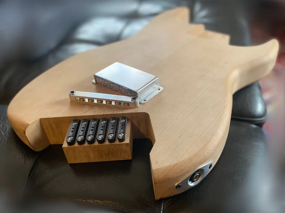 A close-up photo shows the back of an unfinished wooden bass guitar body resting on a black leather couch. Visible…