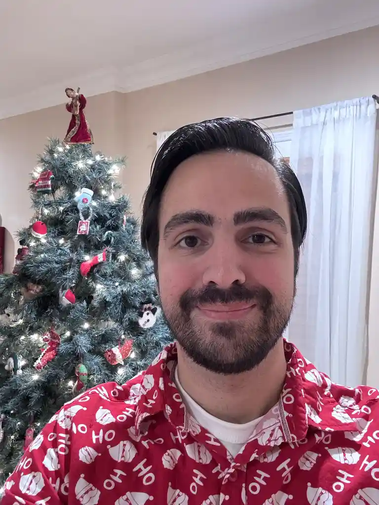A man with dark hair and a beard smiles at the camera while wearing a red pajama shirt covered in white 'ho ho ho' text and…