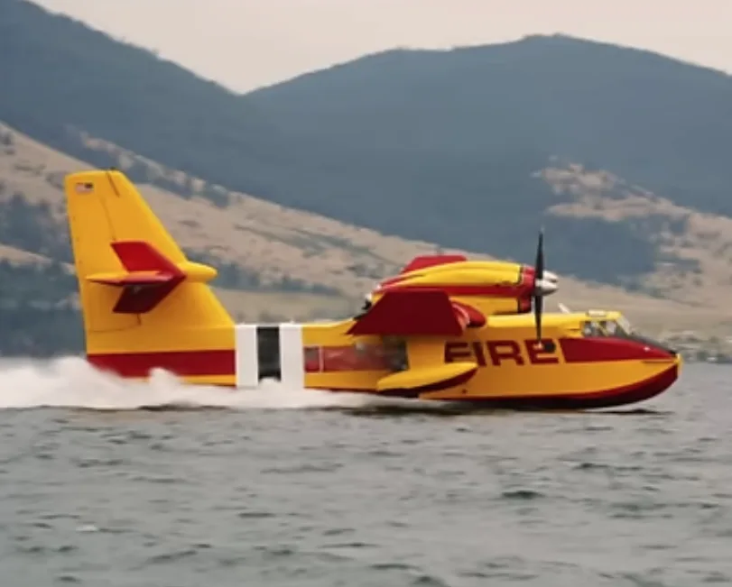 A yellow and red firefighting seaplane with 'FIRE' written on its side is speeding across water, leaving a white wake, with…
