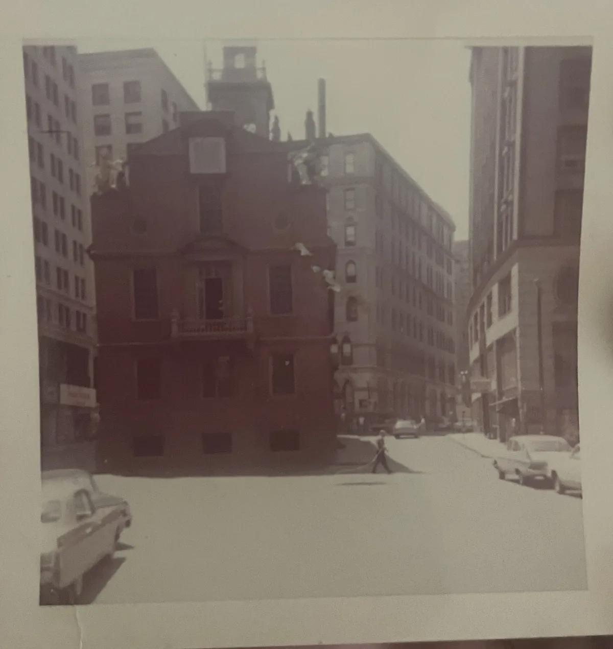 A sepia-toned photograph shows a street scene with vintage cars and tall buildings, including a prominent red-brick building…