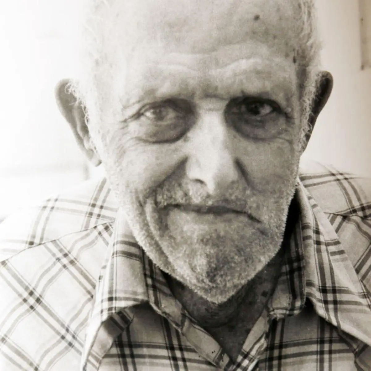 A black and white close-up photo of an elderly man with a weathered face, thinning white hair, and a short beard, wearing a…