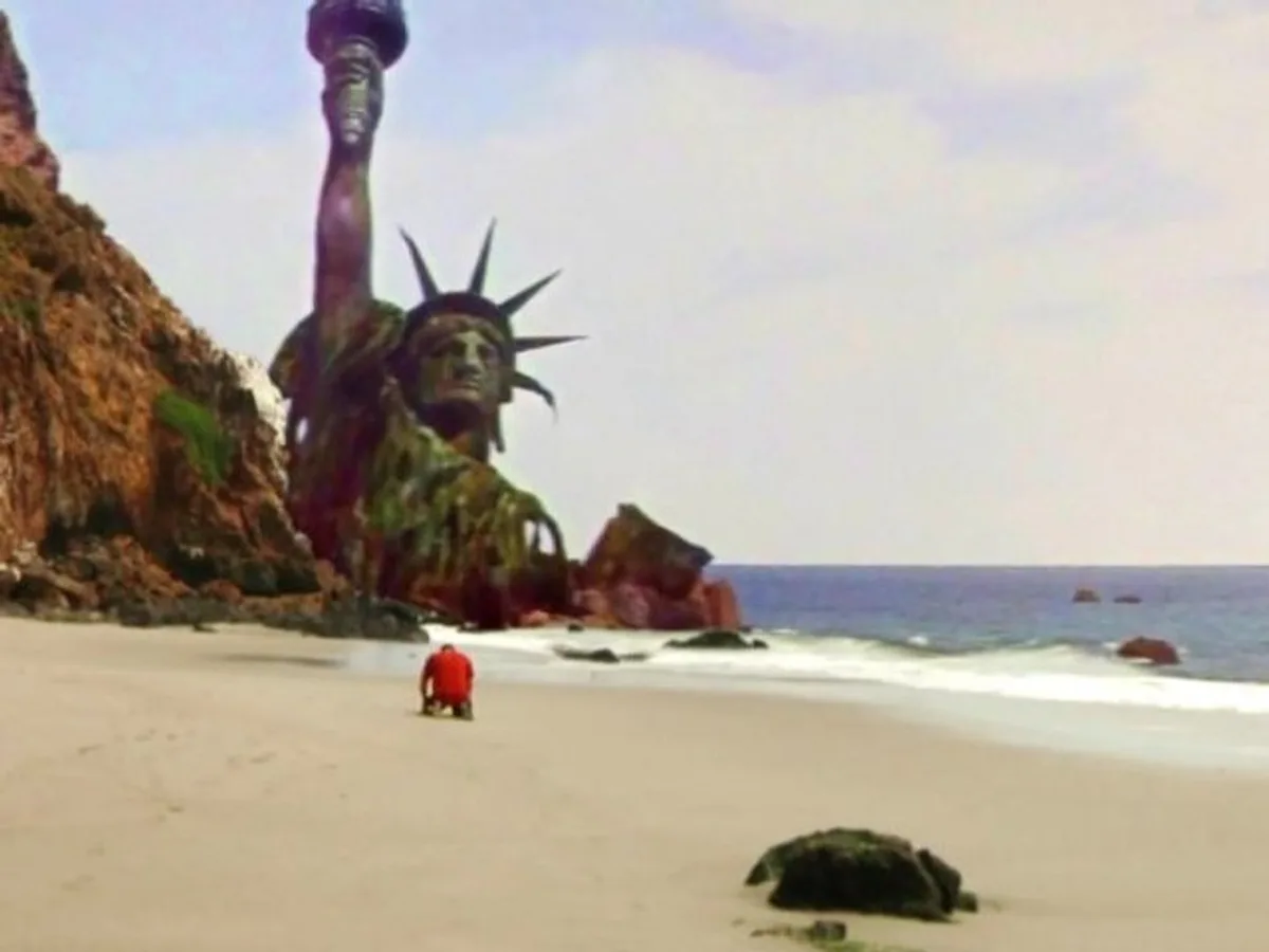 A person in a red shirt kneels on a sandy beach facing a massive, rusted, and partially collapsed Statue of Liberty that…