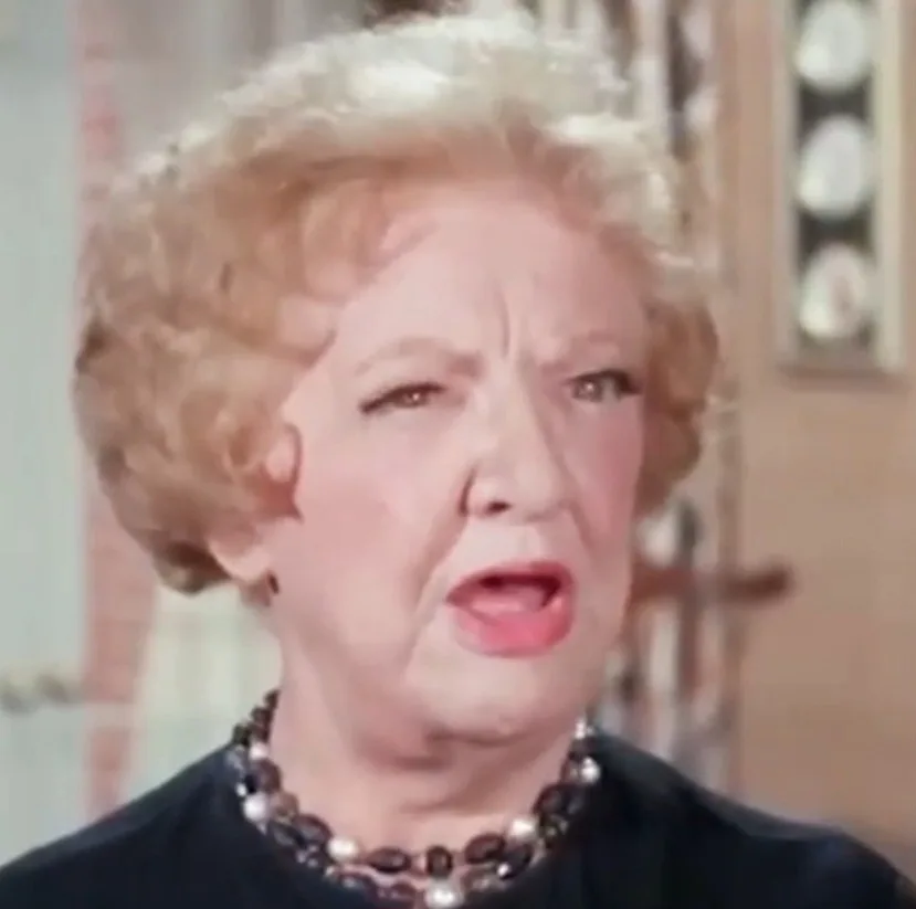 a close-up of an older woman with curly blonde hair, wearing a dark top and a beaded necklace, making a shocked or…