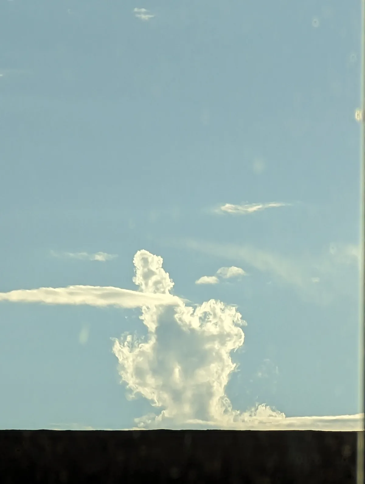 A photograph of a large, puffy white cloud against a pale blue sky, viewed through what appears to be a window with some…