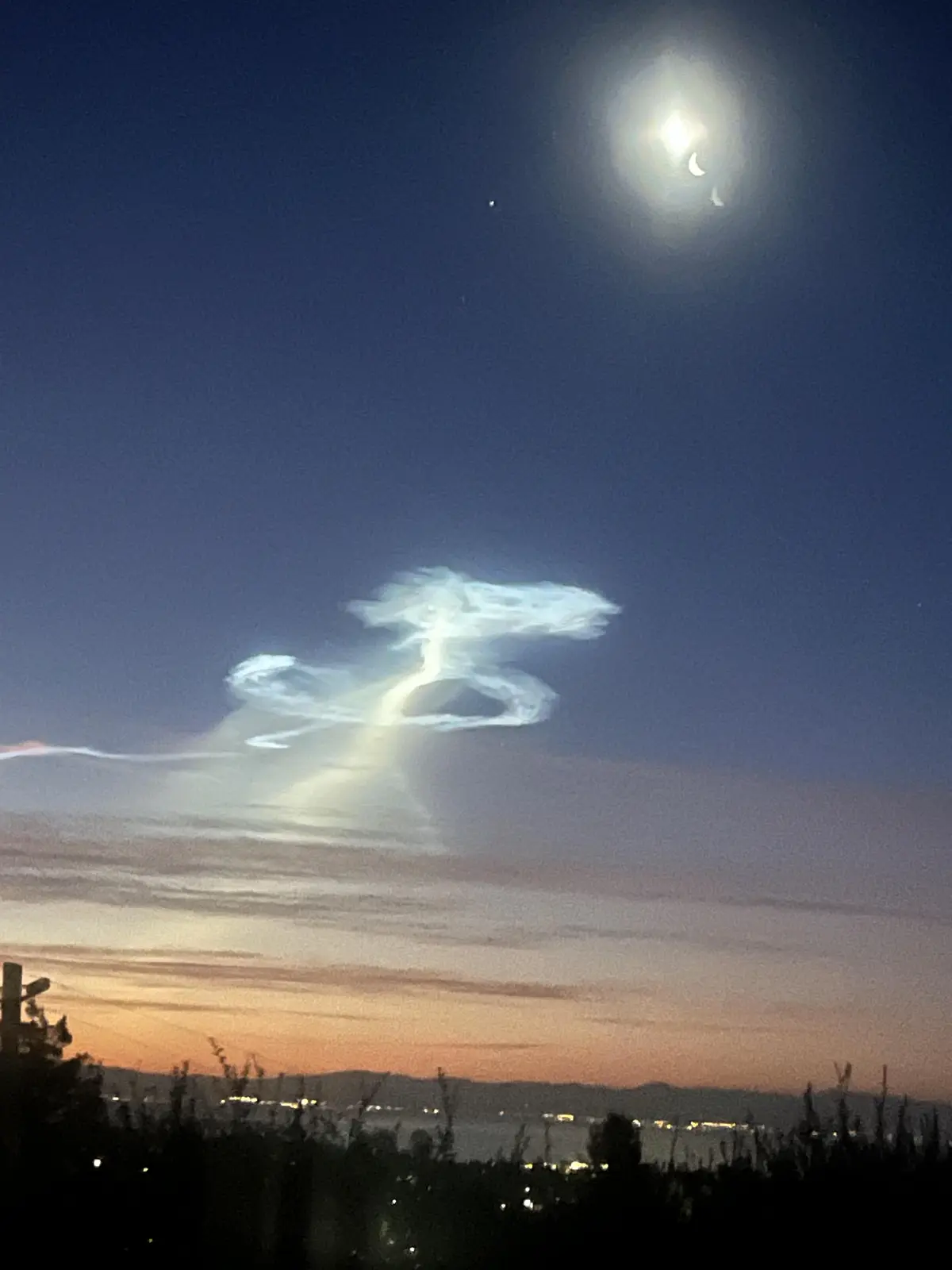 A night sky photo shows a crescent moon partially obscured by clouds, with a large, bright cloud formation below it that…