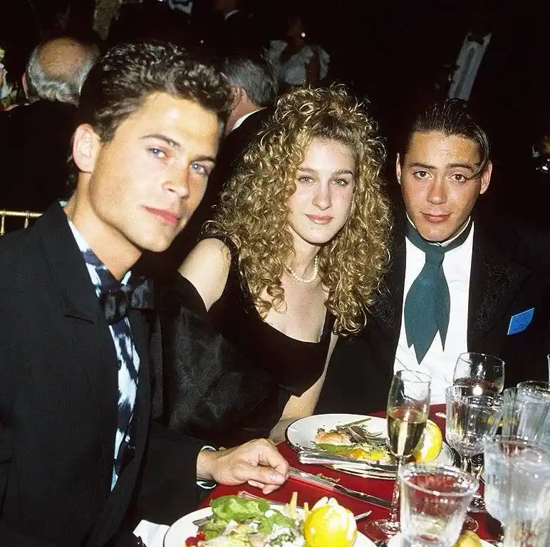 Three people in formal wear sit at a dinner table with plates and glasses; the man on the left and the man on the right both…