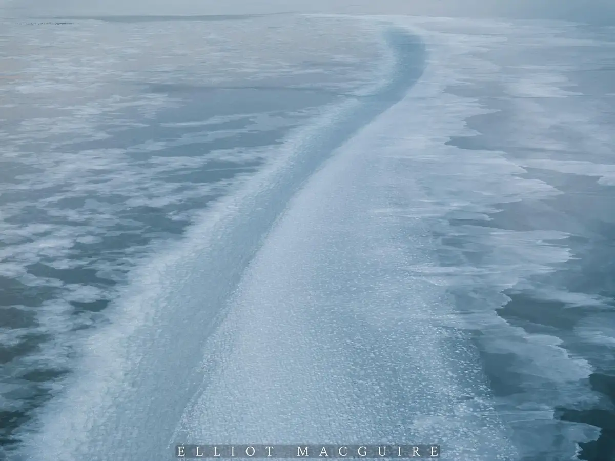 An aerial photograph shows a winding, textured line of ice cutting through a vast, flat expanse of frozen water, with the…
