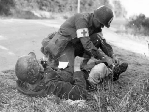 A black-and-white historical photo shows a soldier in a military uniform with a red cross armband kneeling beside another…