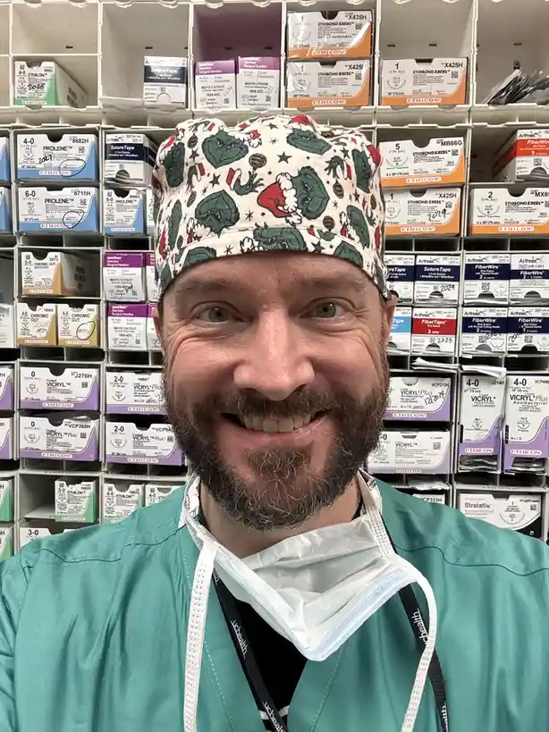 A smiling man with a beard, wearing green surgical scrubs, a surgical mask pulled down under his chin, and a festive…