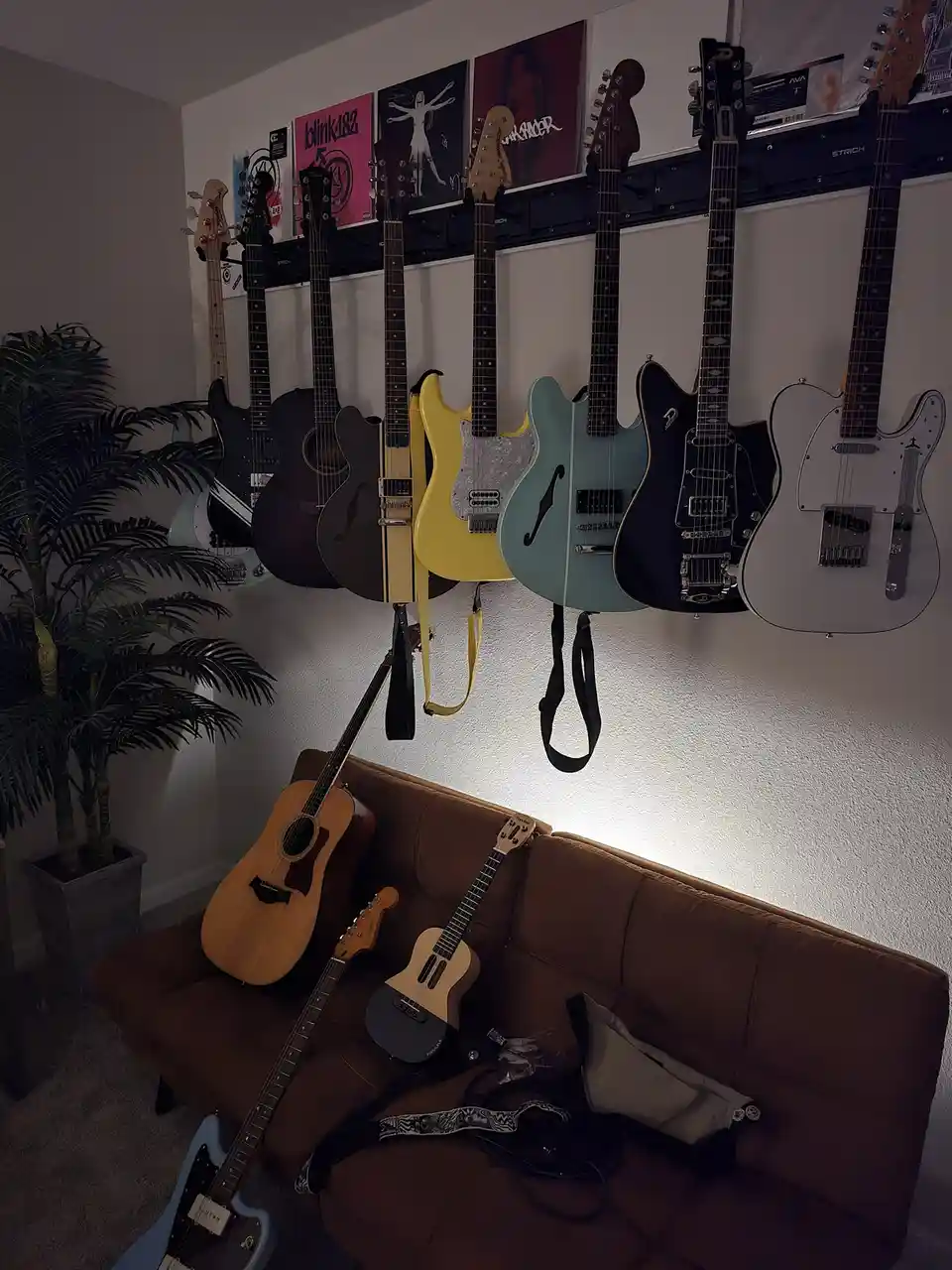 A room with multiple electric and acoustic guitars hanging on a wall rack and resting on a brown couch, with posters of…
