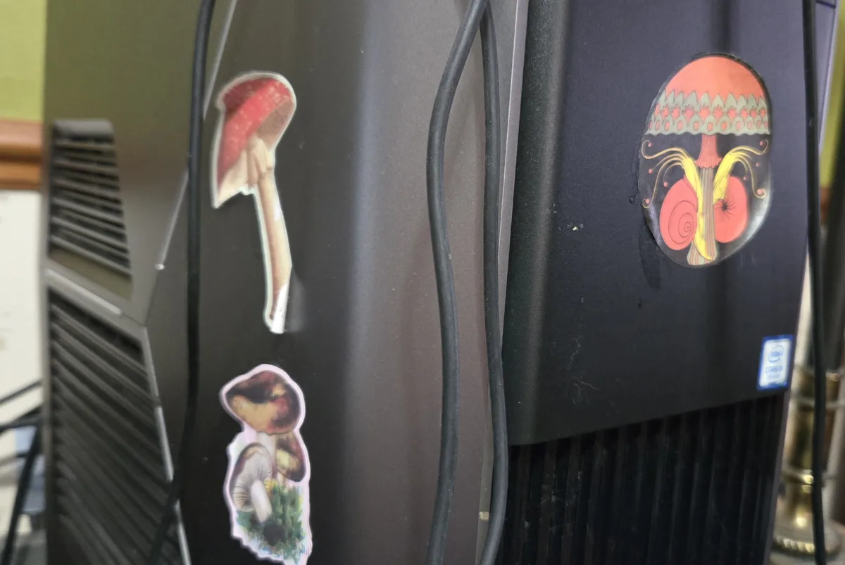 A close-up of a black computer tower decorated with three mushroom-shaped stickers: one red-capped mushroom, one cluster of…
