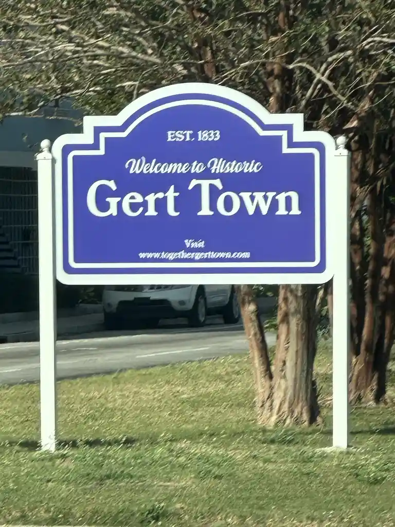A blue and white town sign reads 'EST. 1833 Welcome to Historic Gert Town' with a website below. The sign is mounted on two…