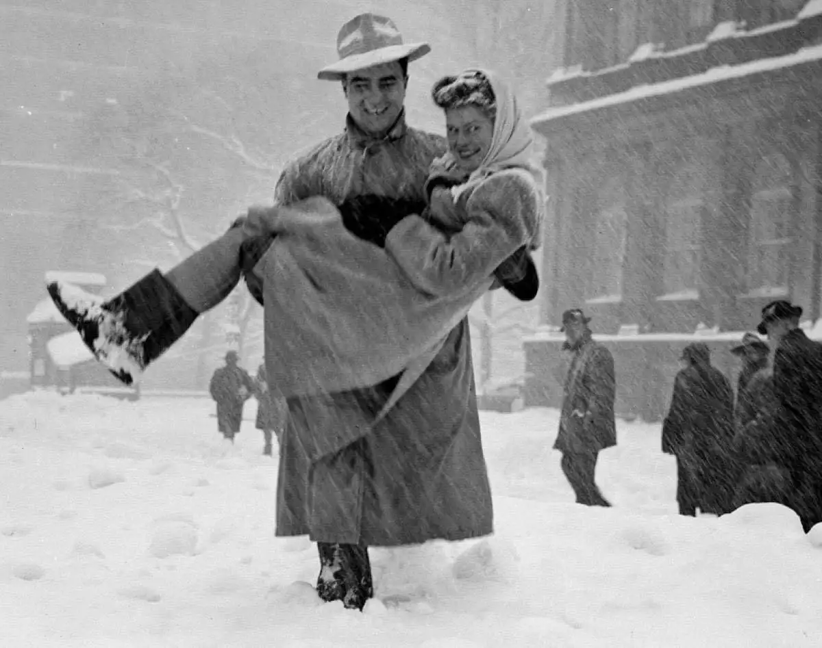 A vintage black-and-white photo shows a man in a hat and coat joyfully carrying a smiling woman in a coat and headscarf…
