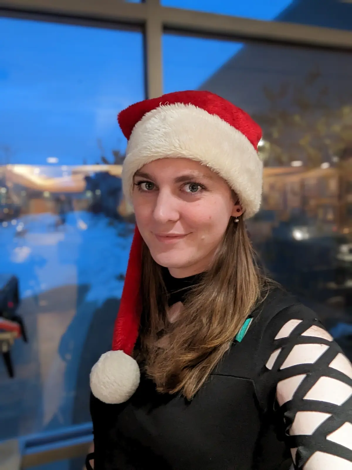 A woman with brown hair and green eyes wears a red and white Santa hat while looking at the camera with a subtle, slightly…