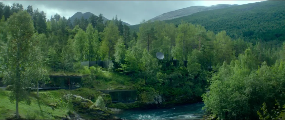 A scenic view of a modern, glass-walled cabin nestled in a lush green forest beside a river, with mountains in the…