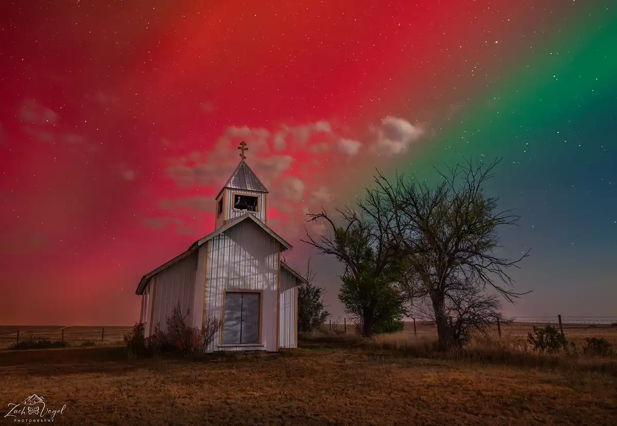 A small, white, wooden church with a cross on its steeple stands in a field under a surreal night sky. The sky is split…
