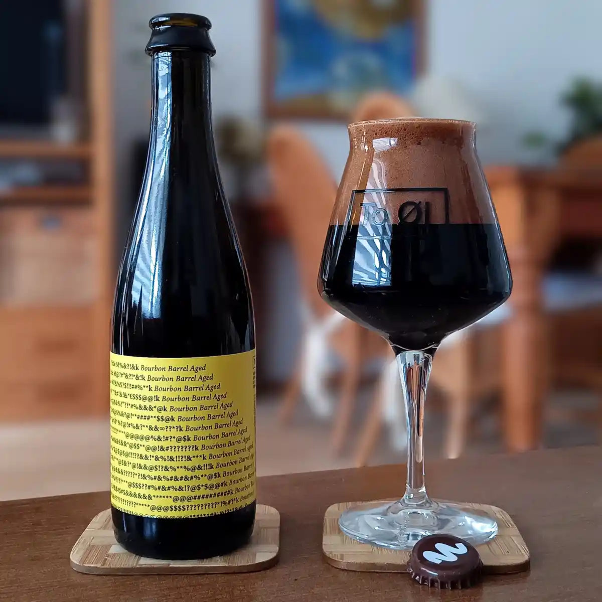 A dark beer bottle and a filled glass sit on coasters, the bottle's label is covered in nonsensical symbols and repeated…