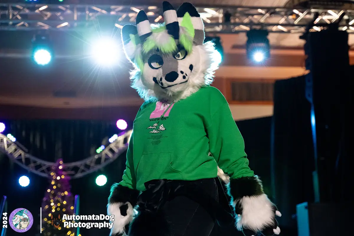A person in a fursuit with a white, grey, and neon green goat-like head with black-tipped horns stands on a stage under…
