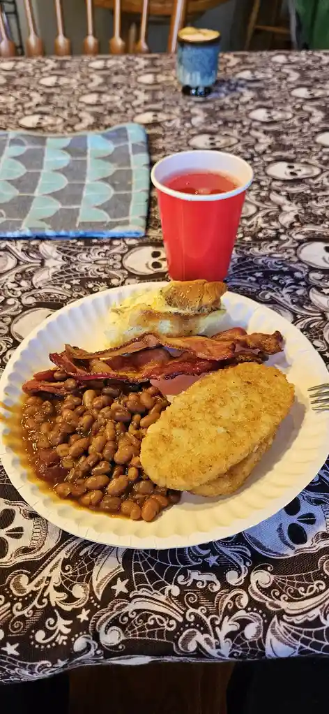 A plate of breakfast food including bacon, baked beans, hash browns, and a piece of bread sits on a table with a…