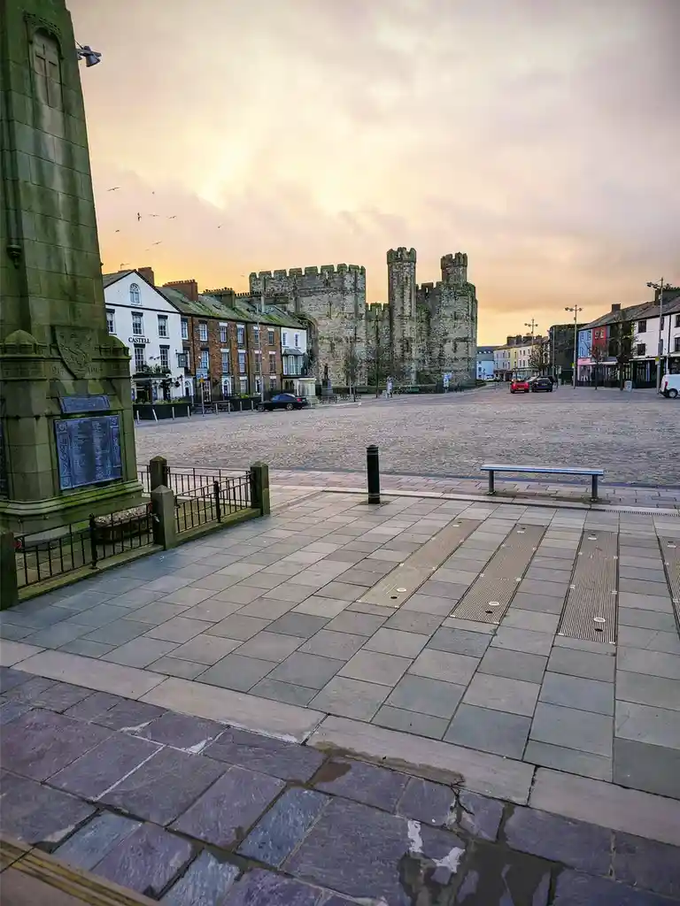 A photo of a quiet town square at dusk, featuring a war memorial in the foreground, cobblestone streets, and a large…