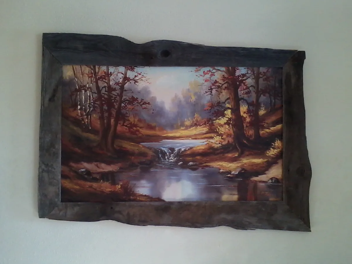 A framed landscape painting of a forest stream with autumn trees hangs on a wall. The rustic wooden frame has rough, uneven…