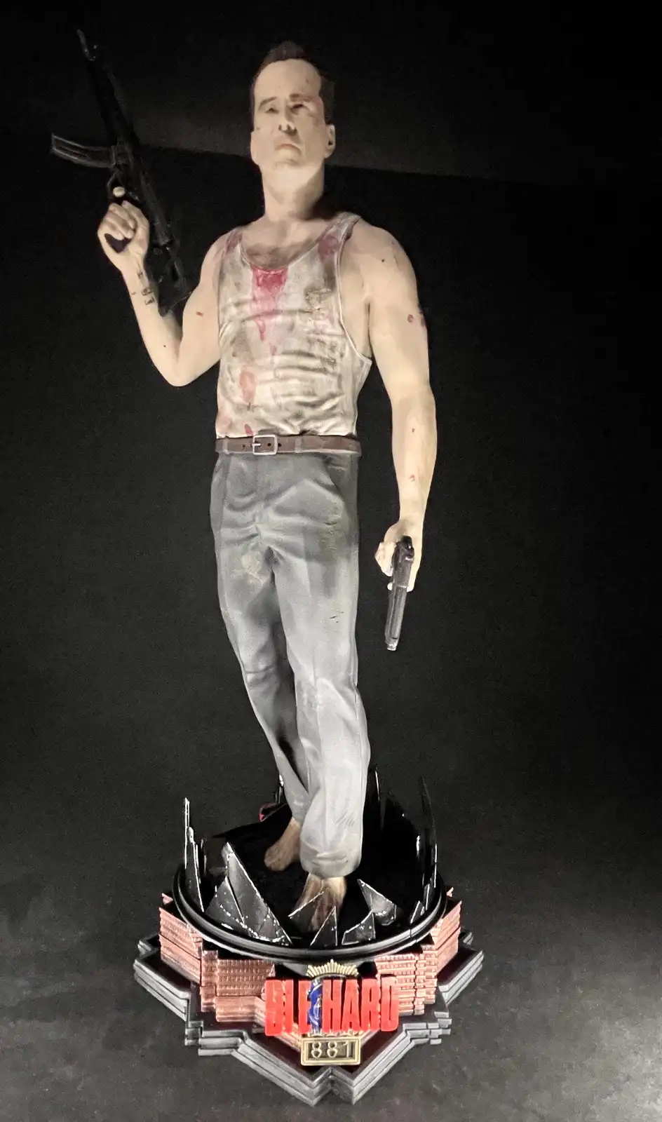 A full-body statue of a man in a blood-stained tank top and pants, holding an assault rifle in one hand and a pistol in the…