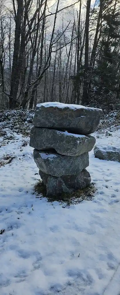 A stack of four large, rough-hewn stones balanced precariously in a snowy forest, with bare trees in the background. The…