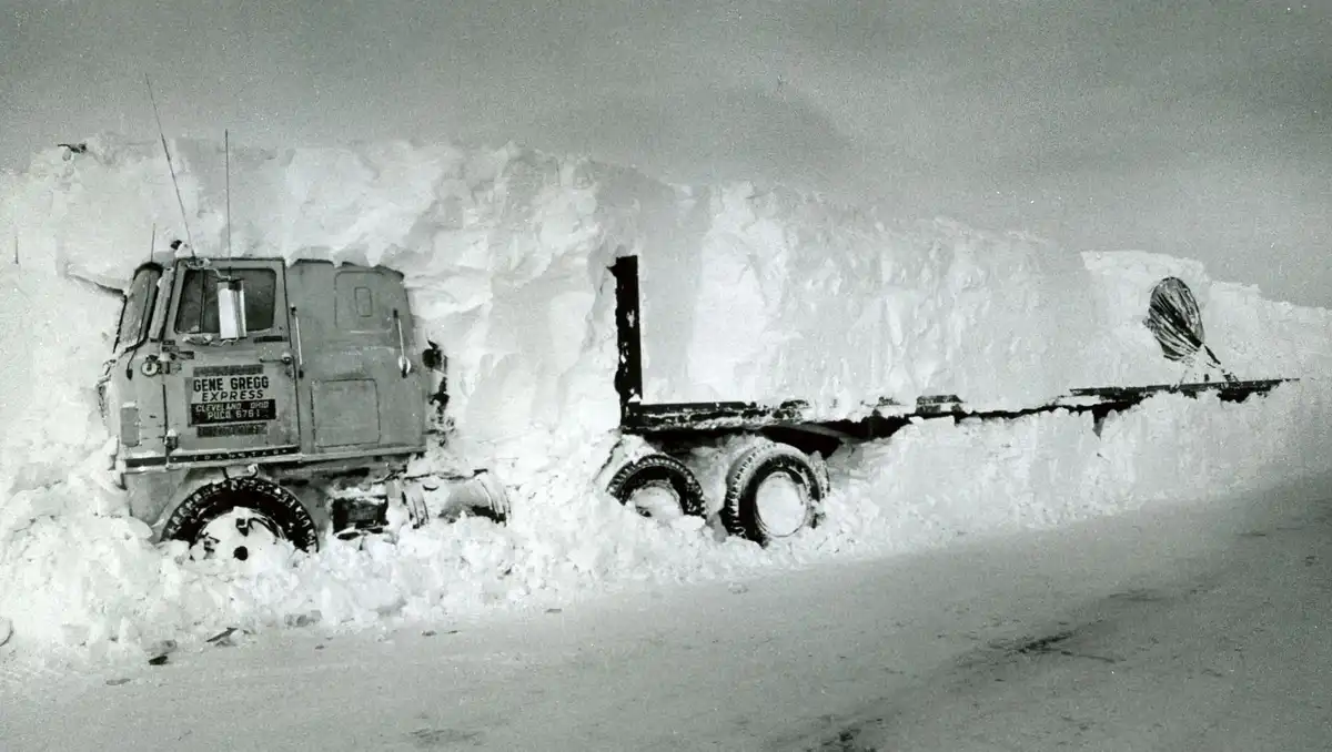 A black and white photo shows a large semi-truck, branded 'GENE GREGG EXPRESS', completely buried in a massive snowdrift…