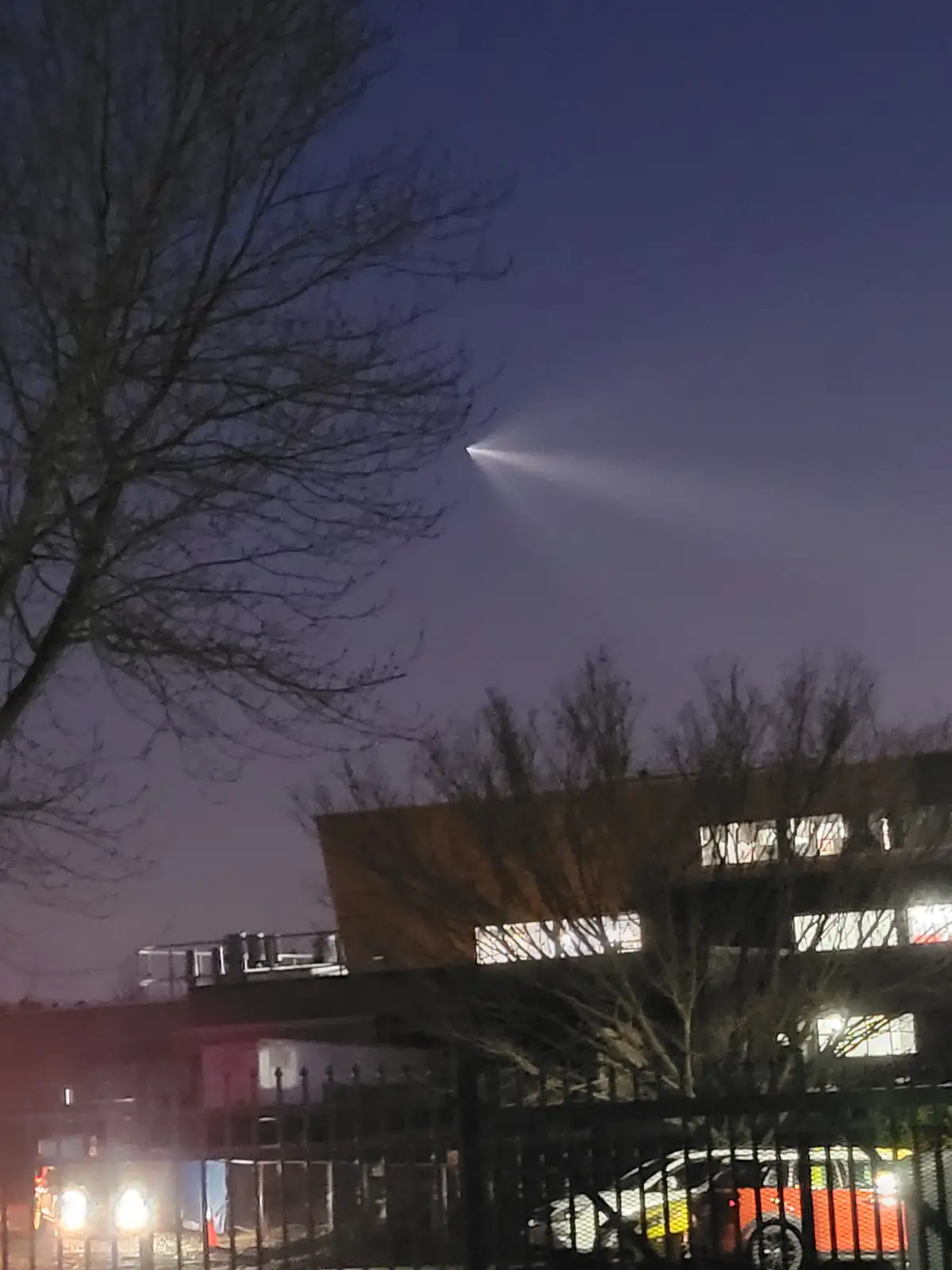 A nighttime photo shows a bright, focused beam of light shooting upward from an unseen source against a dark sky, with bare…
