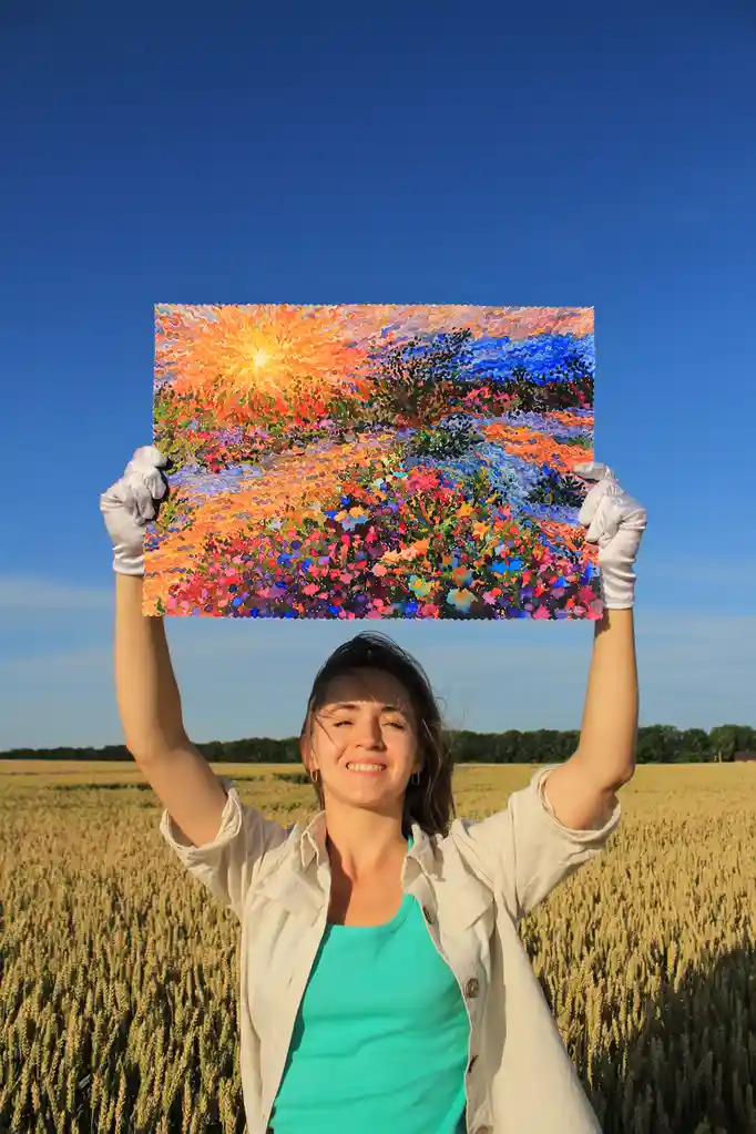 A woman in a wheat field, wearing white gloves, holds up a colorful painting of a sunset over flowers. The painting's…