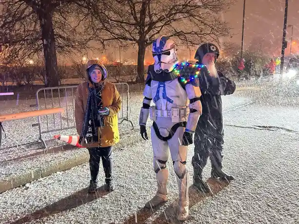 Three people stand outside in heavy snowfall at night. A woman in a brown coat and scarf smiles on the left. In the center…
