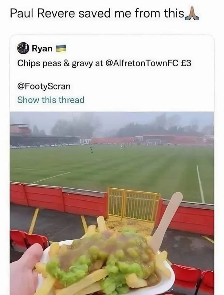 A person holds a plate of chips, peas, and gravy at a football stadium, with a foggy pitch visible in the background. The…