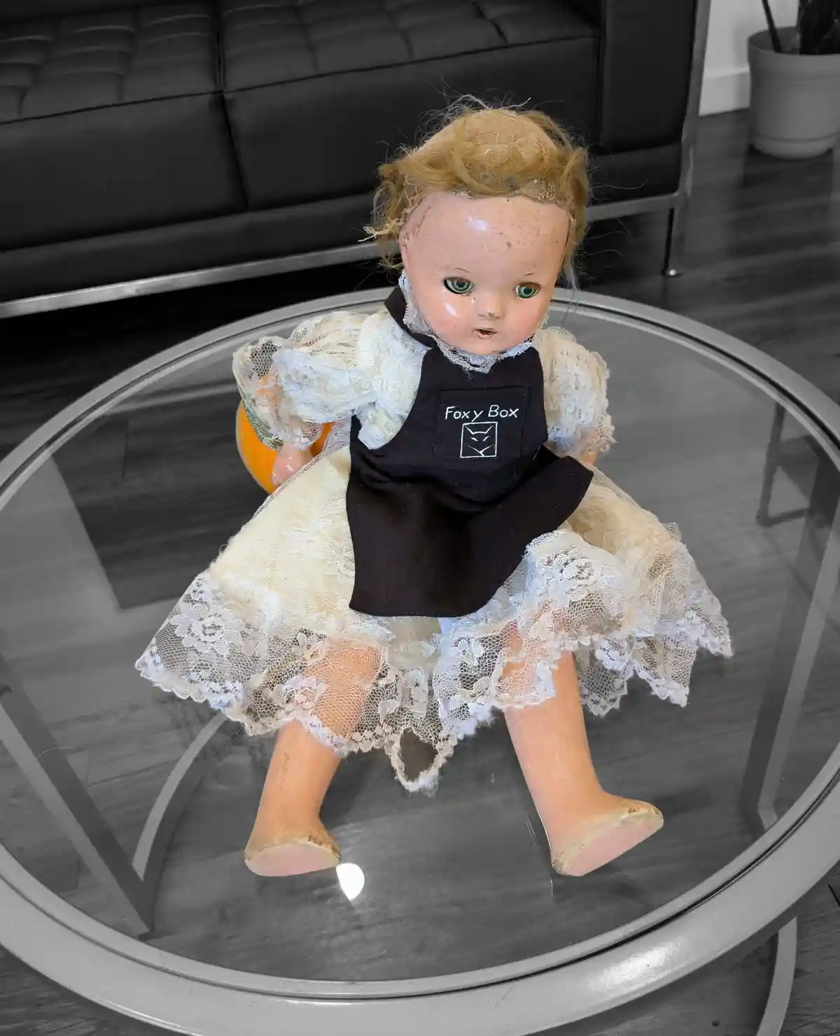 An old, worn doll with blonde hair and green eyes sits on a glass coffee table, wearing a black apron with 'Foxy Box' and a…
