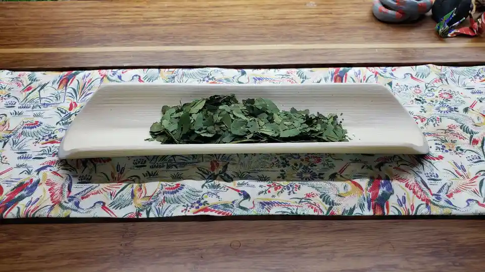 A light-colored wooden tray holds a pile of dried green leaves, resting on a colorful fabric with bird and floral patterns…