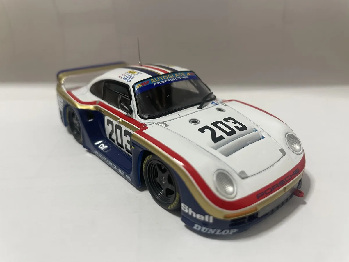 A detailed scale model of a white, blue, and red Porsche 935 race car, number 203, with sponsor logos like Autoglass, Shell…