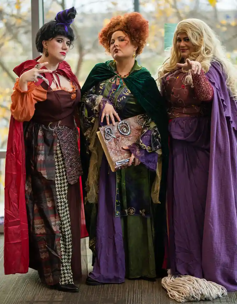 Three women dressed as the Sanderson sisters from Hocus Pocus stand side-by-side indoors, each striking a confident pose…